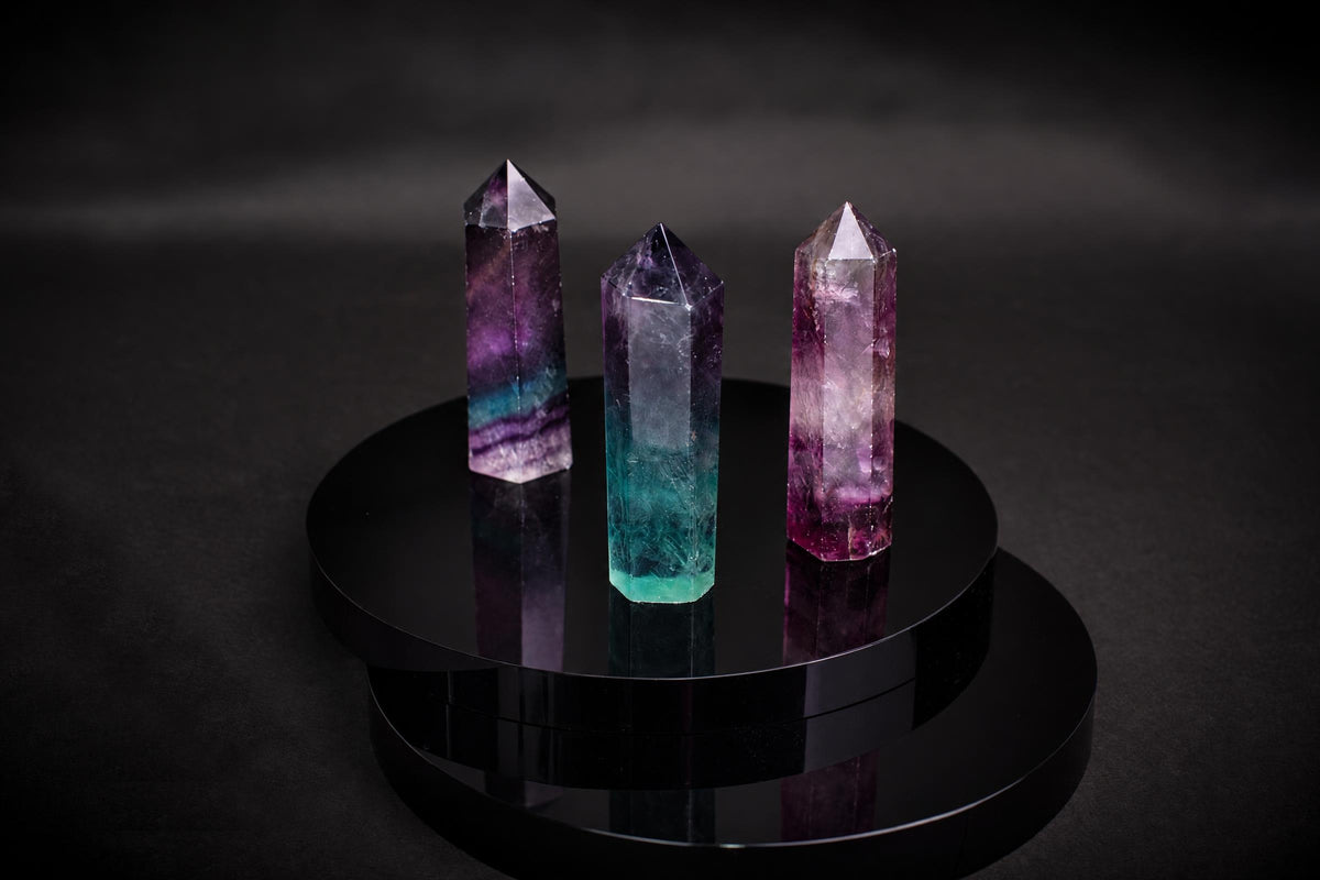 Three fluorite crystal points in purple, teal and pink stand on a dark reflective surface.