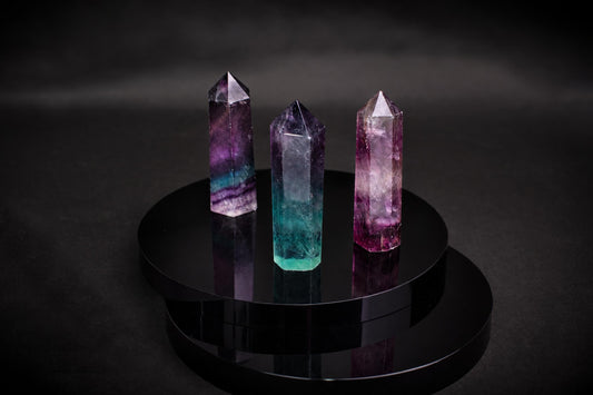 Three fluorite crystal points in purple, teal and pink stand on a dark reflective surface.