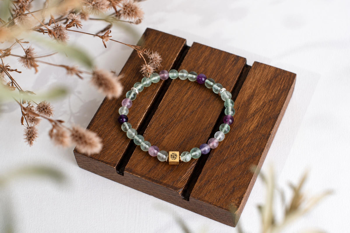 A bracelet of pale-green and purple beads with small metallic spacers rests on a wooden board beside dried stems.