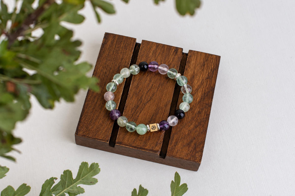 A beaded bracelet with green, purple, and black stones rests on a dark wooden stand against a white surface.