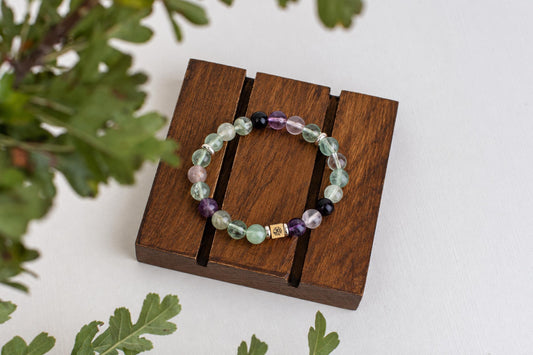 A beaded bracelet with green, purple, and black stones rests on a dark wooden stand against a white surface.