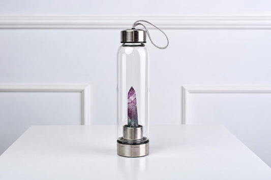 A clear glass water bottle with a silver-toned lid and base, containing a purple fluorite crystal on a white surface.