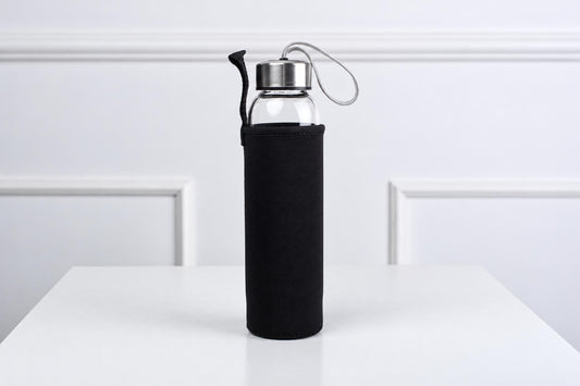 A clear glass water bottle with a silver-toned lid and black fabric sleeve, standing on a white surface.