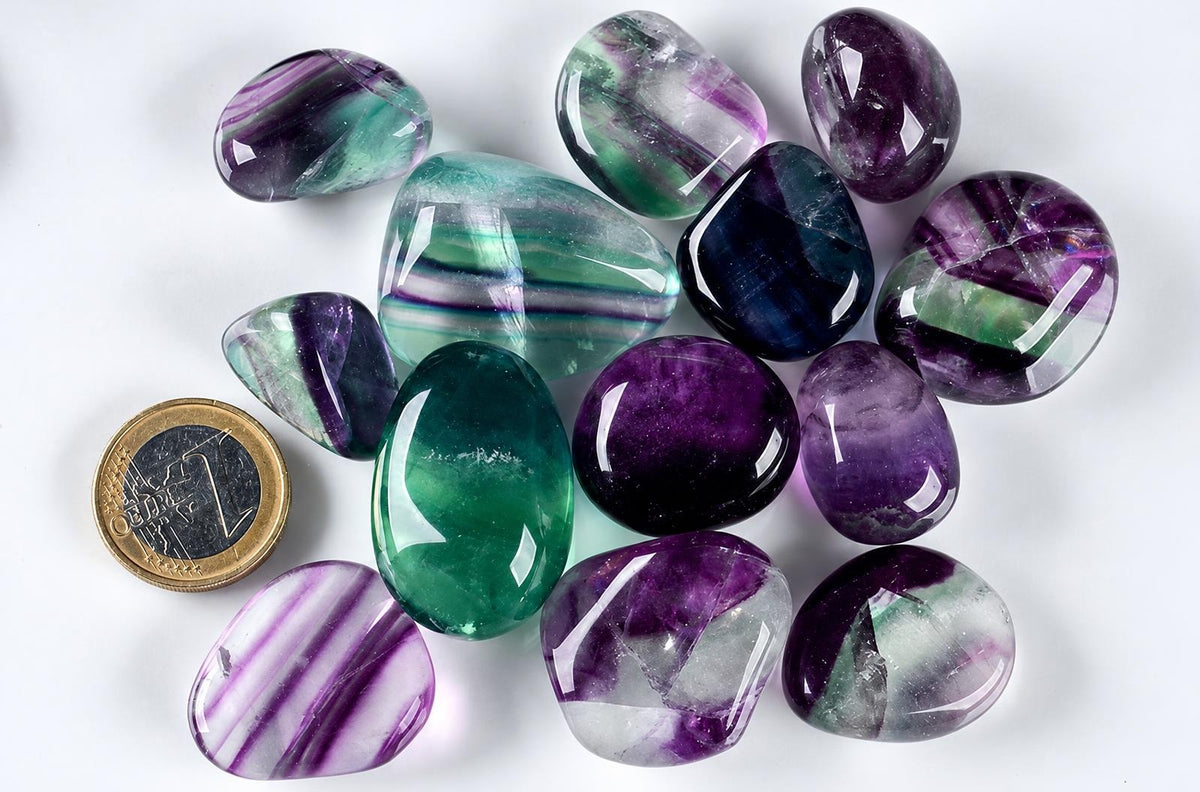A collection of polished fluorite tumbled stones in purple, green, and blue hues on a white surface with a Euro coin for scale.