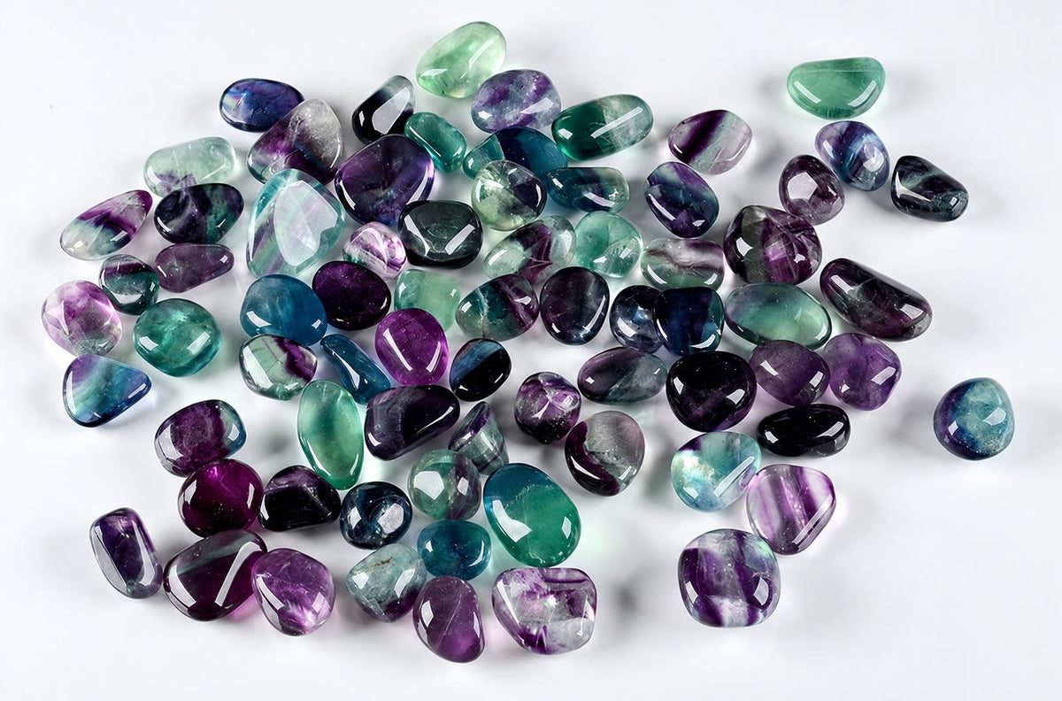 A pile of polished fluorite tumbled stones in green, purple, blue and black on a white surface.