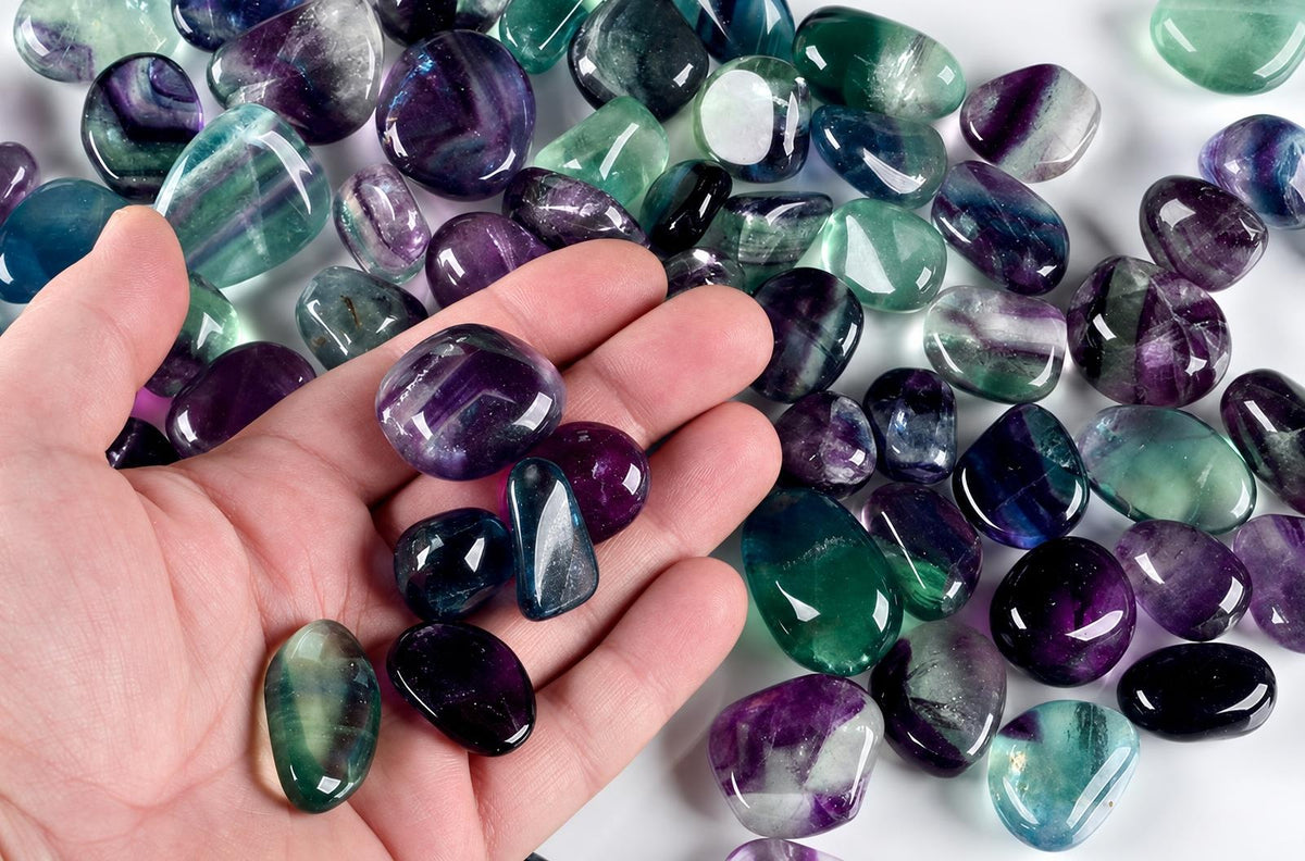 A hand holds polished fluorite tumbled stones showing purple, green and blue bands on a white surface.