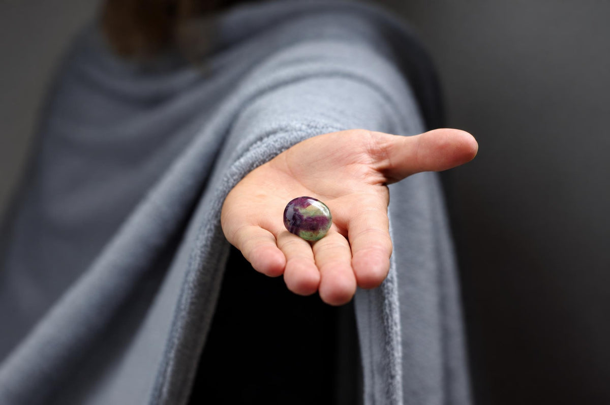 A person in a grey robe holds a polished, oval stone with purple and green swirls in their open palm against a dark background.