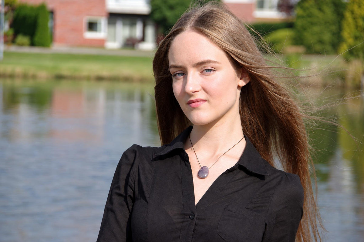 A woman wears a black shirt and a pendant with a dark purple, oval stone on a thin cord against a blurred water background.