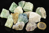 A collection of raw fluorite crystals in green, blue, white and yellow tones on a dark surface with a one euro coin for scale.