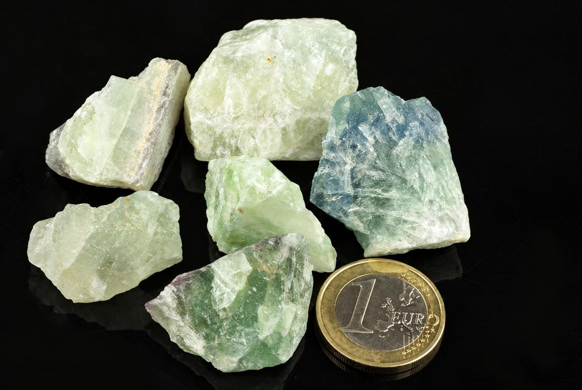 Six raw fluorite crystals in pale green, blue-green and white tones on a dark surface with a one-euro coin for scale.