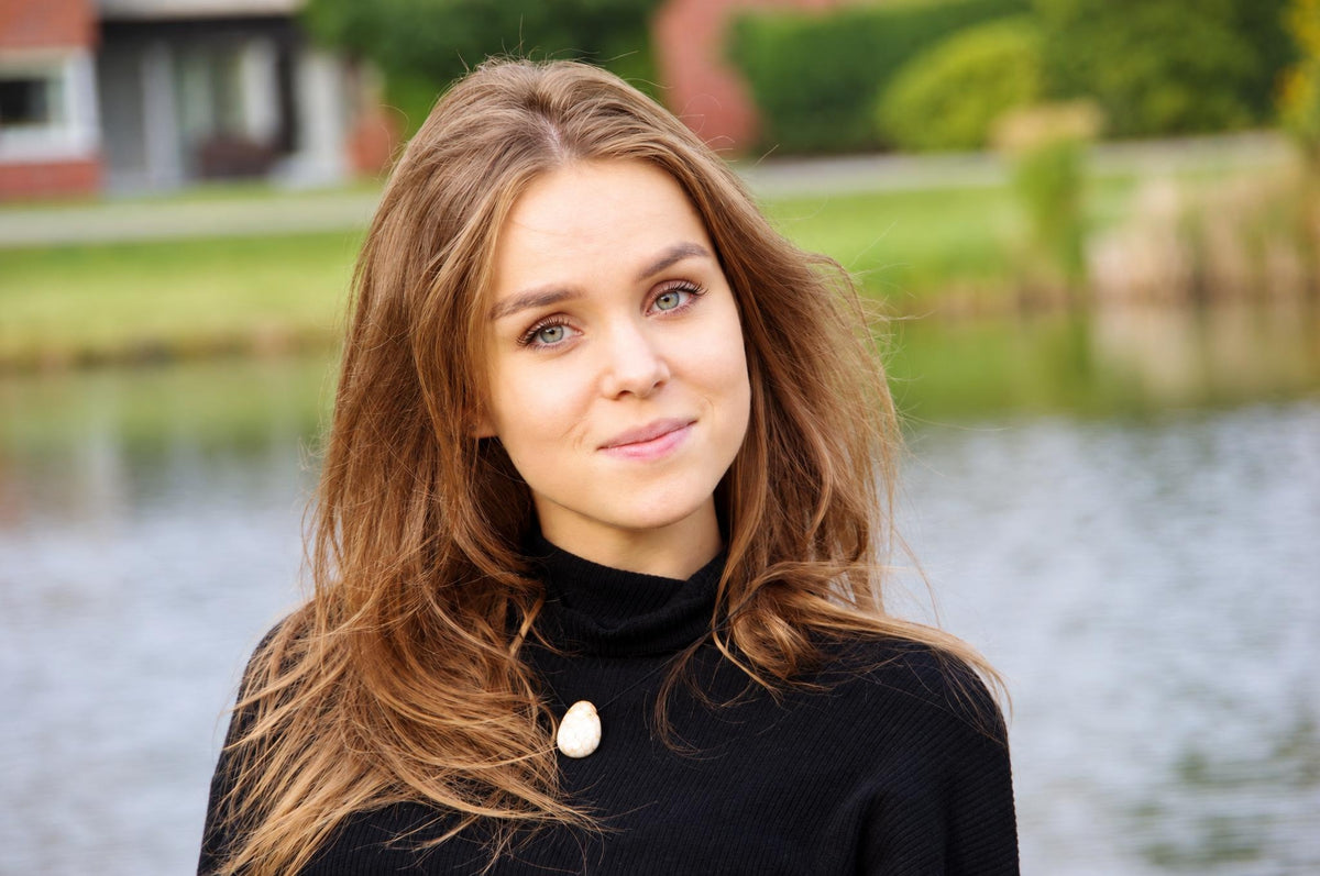 A woman with long brown hair wears a black turtleneck and a white oval pendant on a dark surface near water.