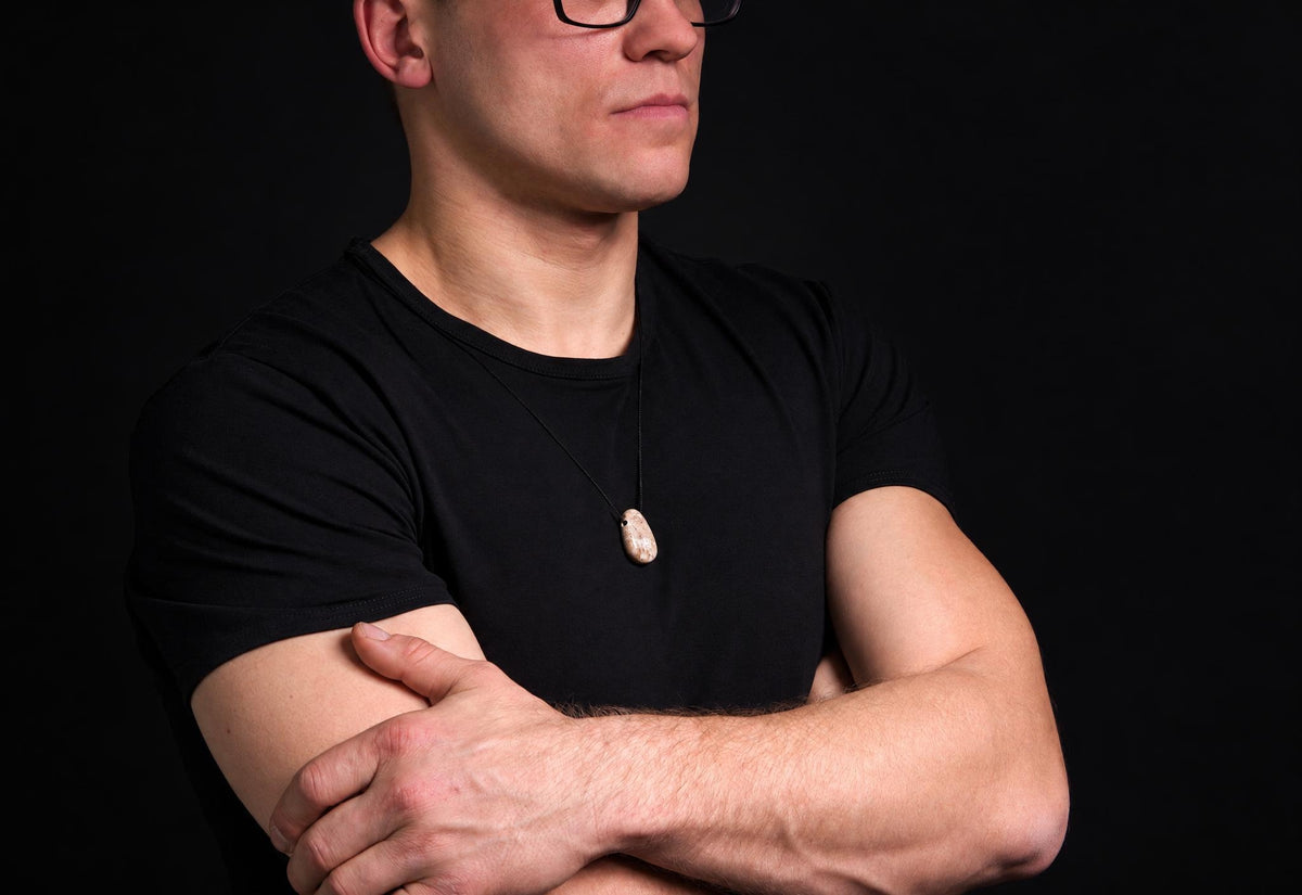 A man in a black t-shirt wears a necklace with an oval, light-colored pendant on a dark surface.
