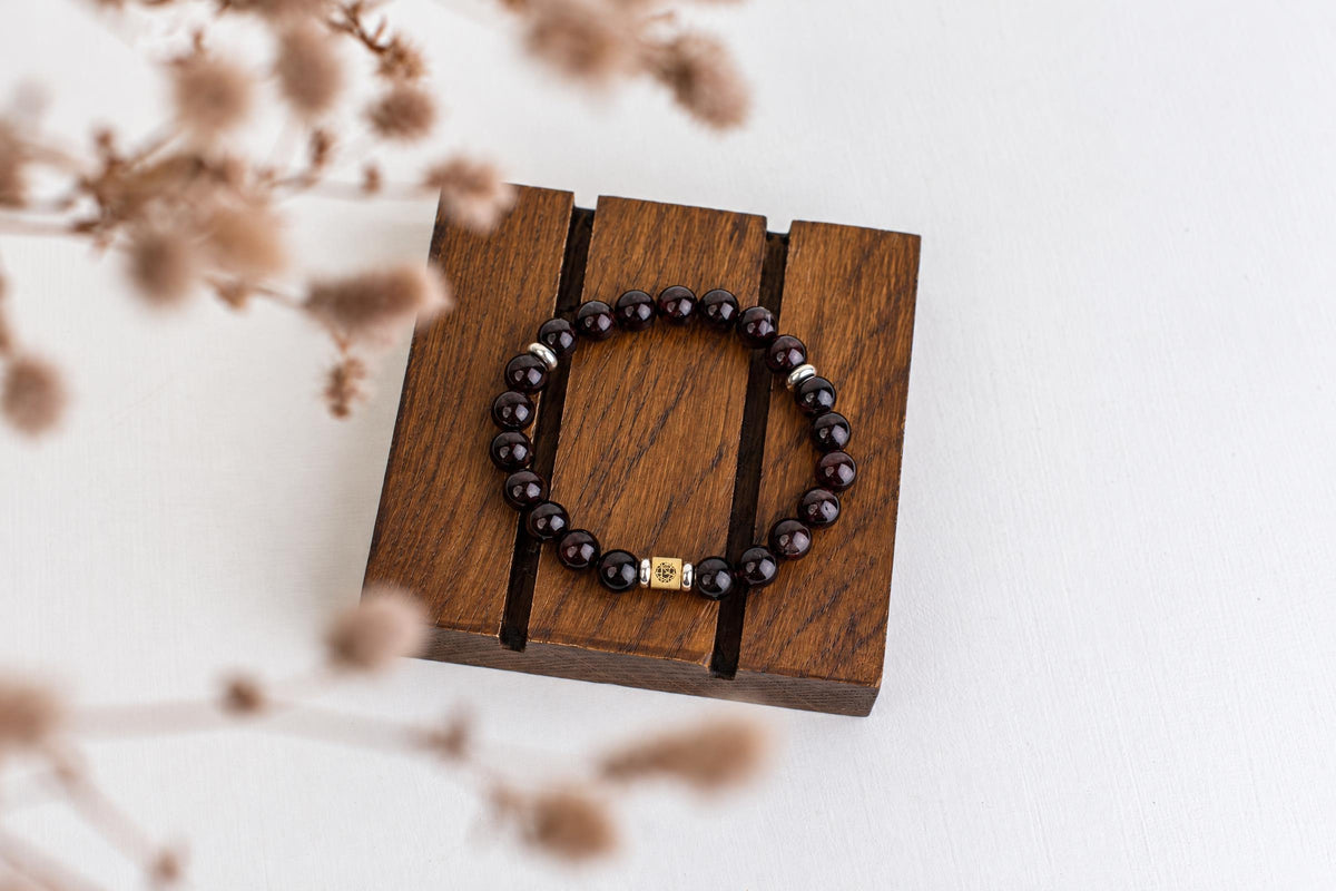 A bracelet with dark red beads and a gold-toned central charm rests on a wooden stand, surrounded by dried flowers.