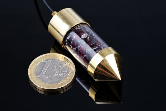 A brass pendulum with a clear tube containing red garnet stones, resting on a dark surface next to a one-euro coin.