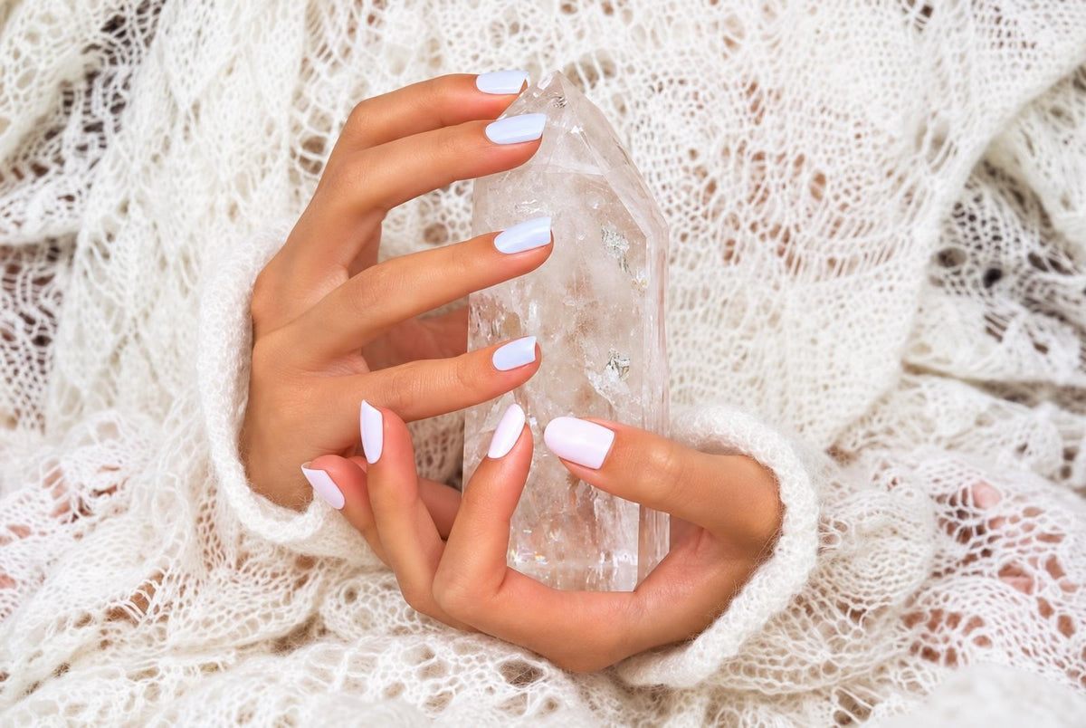 Two hands with pale pink nails hold a clear, pointed crystal on a white knitted fabric.