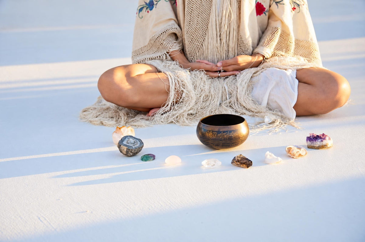 A person in a cream fringed poncho sits cross-legged on a pale surface with a bronze singing bowl and assorted crystals arranged before them.