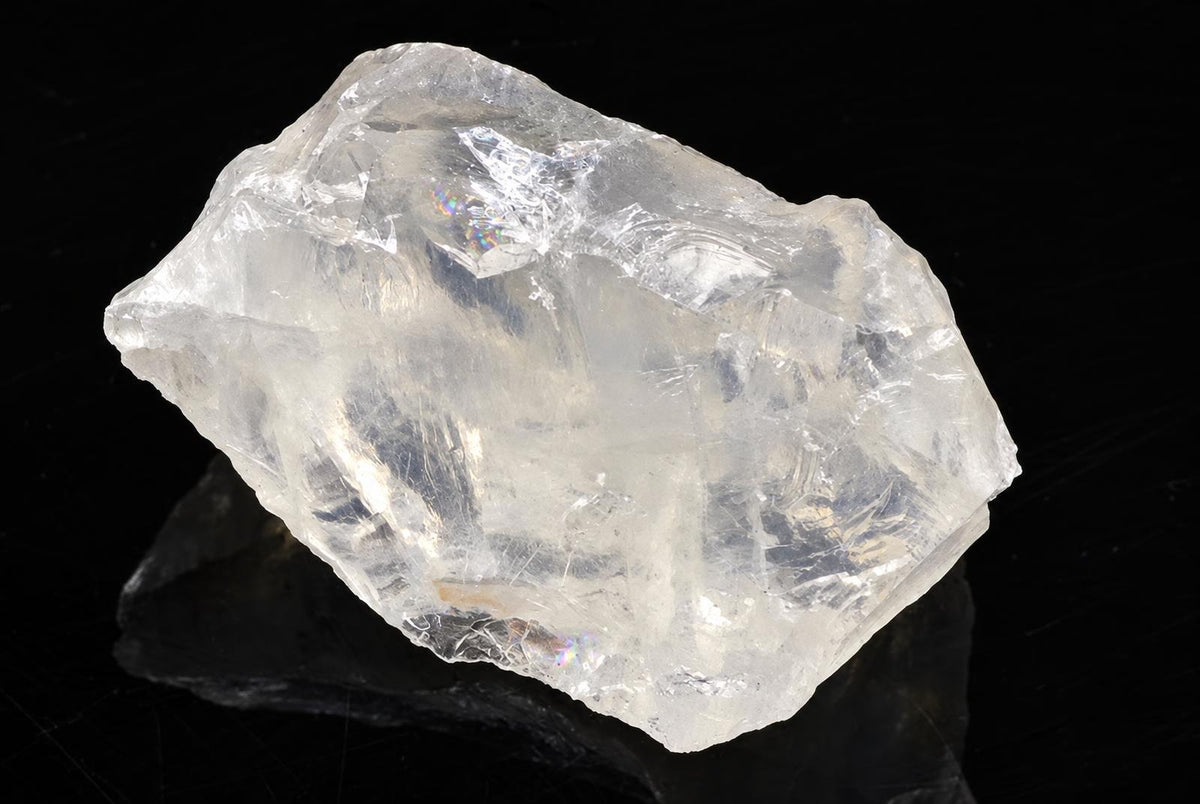 A large, clear, translucent crystal with a rough, angular shape rests on a dark surface against a black background.