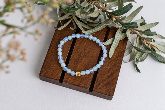 A bracelet of pale-blue beads with small metallic spacers rests on a wooden board beneath leafy branches.