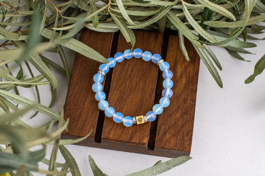 A bracelet with round, light blue beads and a gold-toned square charm rests on a dark wooden surface surrounded by green leaves.