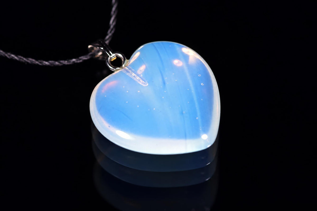 A heart-shaped glass opal pendant with blue and white swirls hangs from a dark cord against a black background.