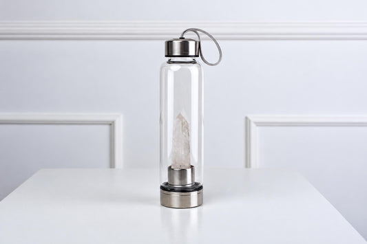 A clear glass water bottle with a silver-toned lid and base, containing a tall, pale quartz crystal inside, standing on a white surface.