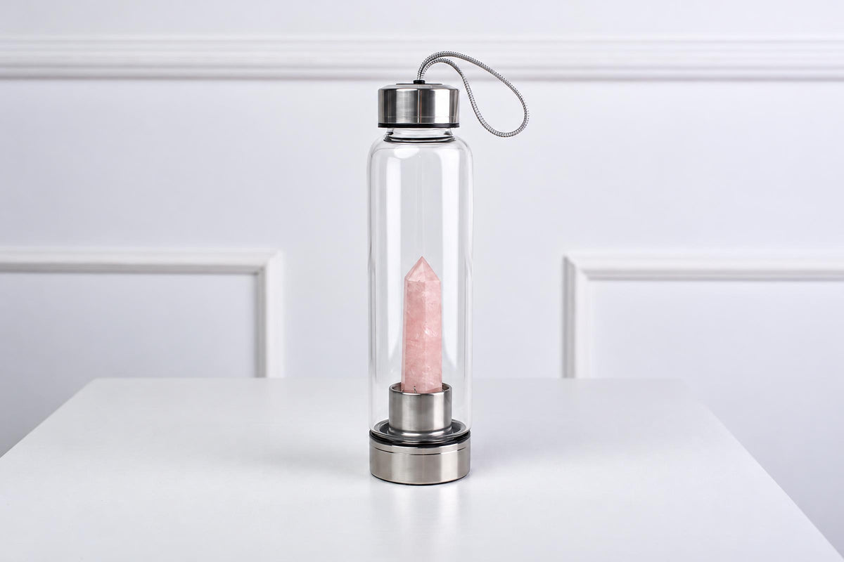 A clear glass water bottle with a silver-toned lid and base, containing a pink rose quartz crystal on a white surface.