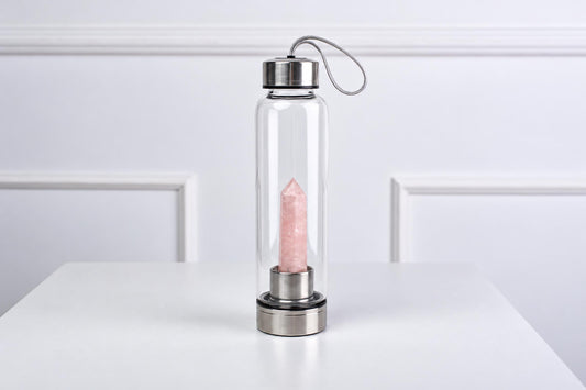 A clear glass water bottle with a silver-toned lid and base, containing a pink rose quartz crystal on a white surface.