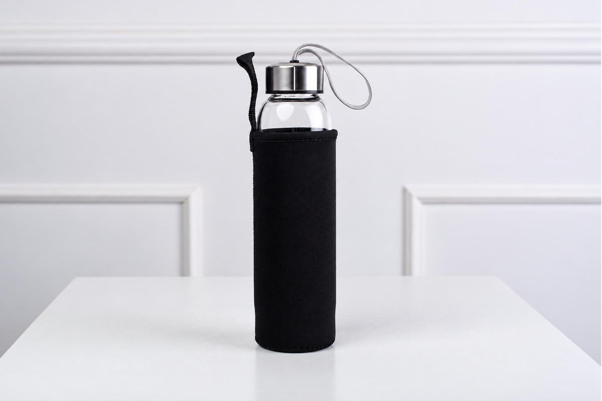 A glass water bottle with a silver-toned lid and black sleeve stands on a white surface against a white wall.