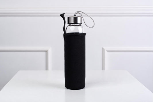 A glass water bottle with a silver-toned lid and black sleeve stands on a white surface against a white wall.