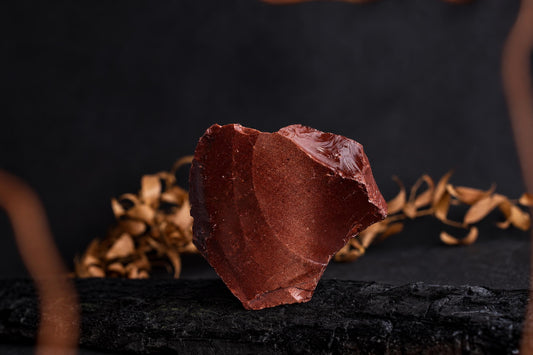 A reddish-brown, irregularly shaped chunk of goldstone rests on a dark surface with dried leaves in the background.
