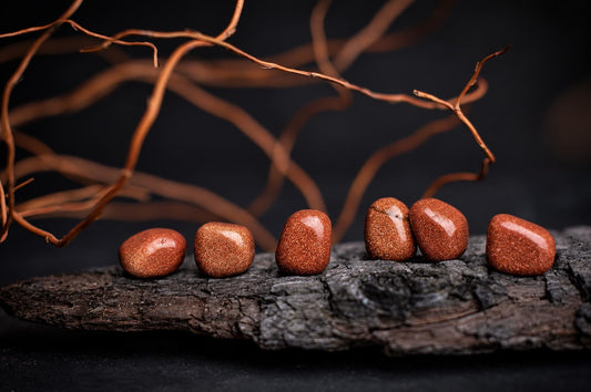 Six polished, reddish-brown Goldstone stones with a sparkly texture are arranged on a dark wooden surface against a black background.