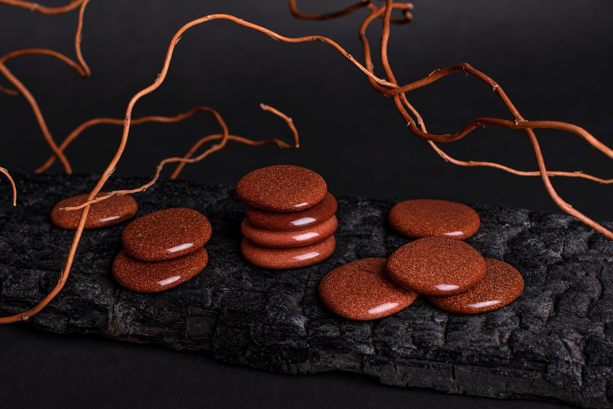 Polished, round goldstone palmstones in reddish-brown with a glittery texture are arranged on a dark surface.