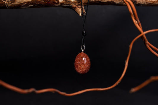 A polished, oval-shaped goldstone pendant with a sparkly orange-brown surface hangs from a black cord on a dark background.