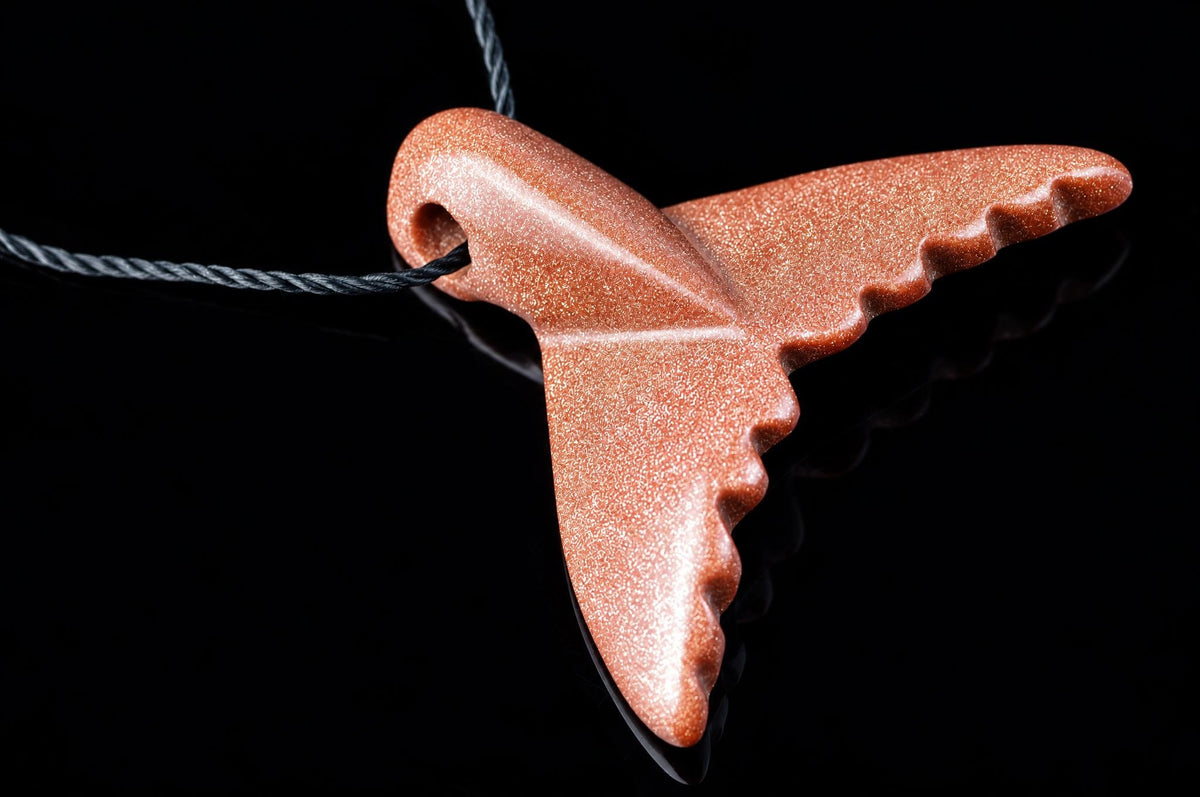 A goldstone pendant shaped like a dolphin tail hangs from a dark cord against a black reflective surface.