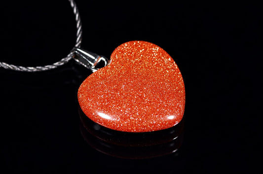 A heart-shaped pendant with a glittering orange-red surface, attached to a silver-toned cord against a dark reflective background.