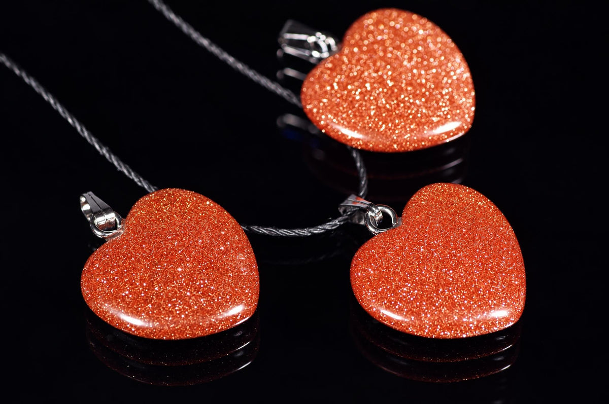 Sparkling reddish-brown heart-shaped goldstone pendants on black cords lie on a dark reflective surface.