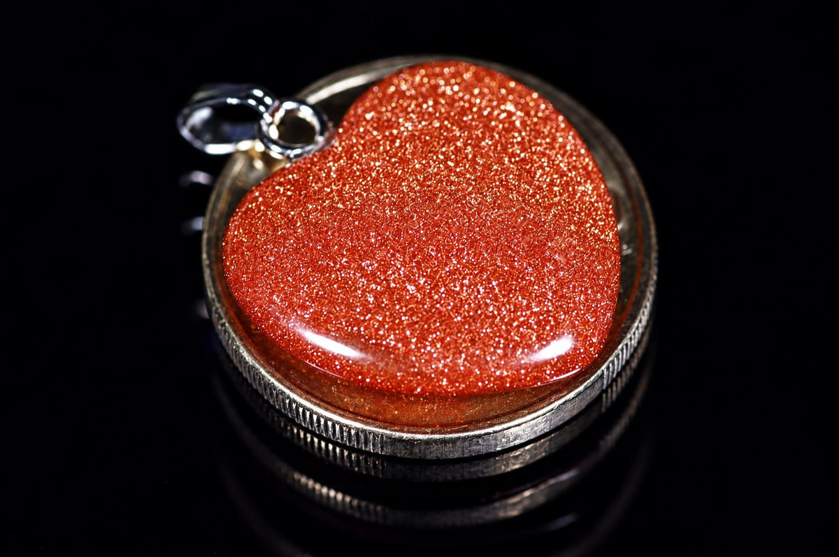 A silver-toned pendant with a red glittery heart-shaped stone set in a circular bezel on a dark reflective surface.