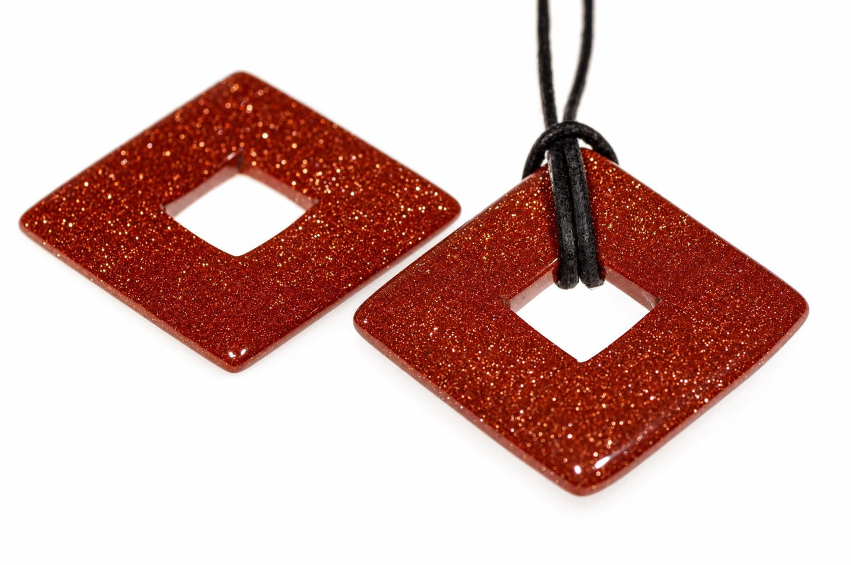 Two square, glittery red pendants with a central hole; one hangs from a black cord on a white surface.