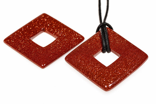 Two square, glittery red pendants with a central hole; one hangs from a black cord on a white surface.