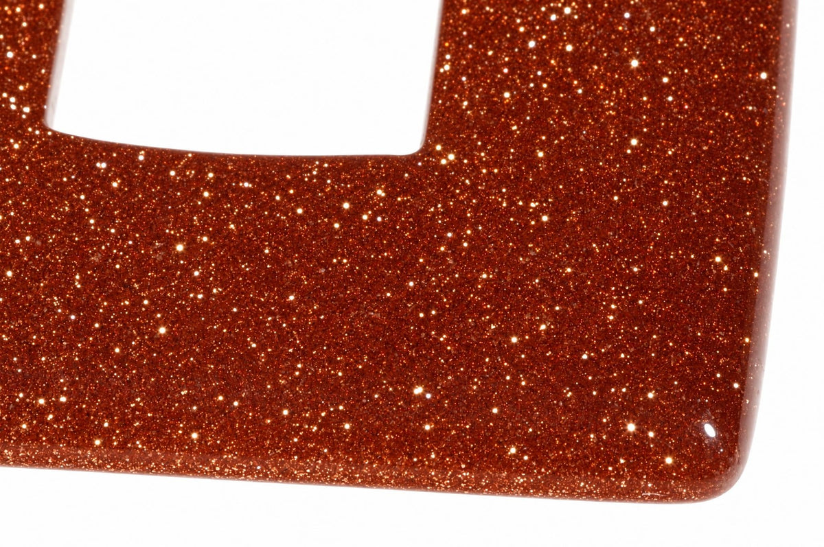 A close-up of a square, gold-coloured pendant with a glittery texture and a cutout corner on a white surface.