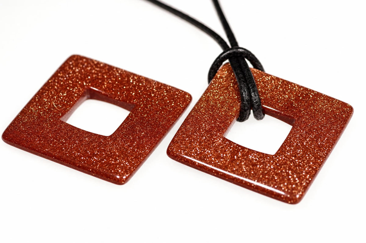 Two square, glittery reddish-brown pendants with central cutouts on a white surface; one has a black cord.