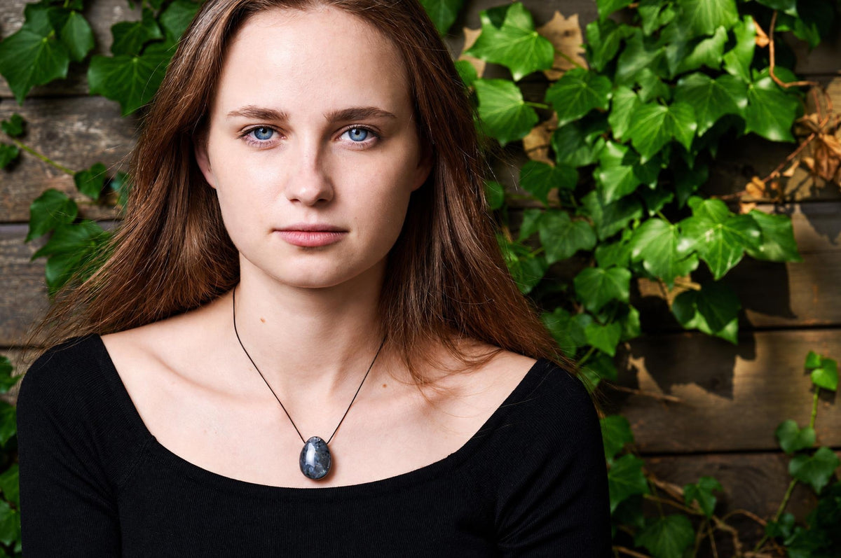 A woman wears a black top and a pendant with an oval, grey-blue stone on a thin cord against a wooden wall with ivy.