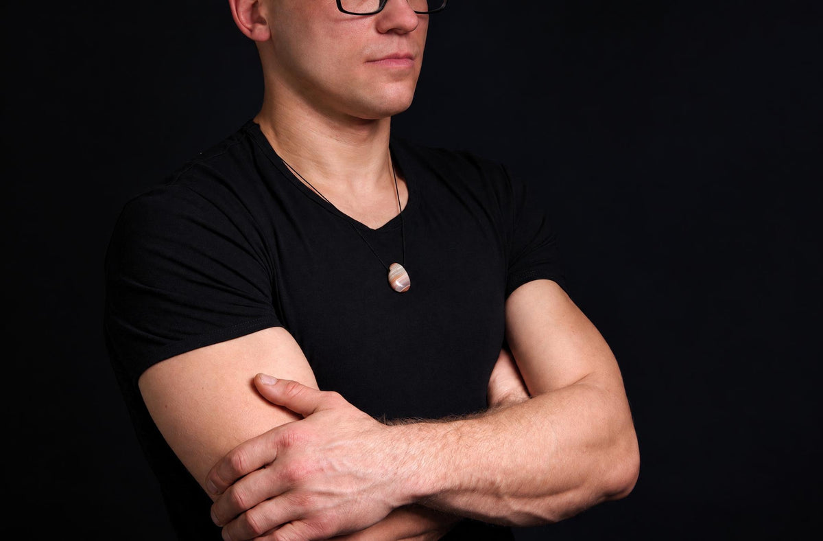 A man in a black t-shirt wears a pendant with a smooth, oval stone showing bands of cream and brown on a dark surface.