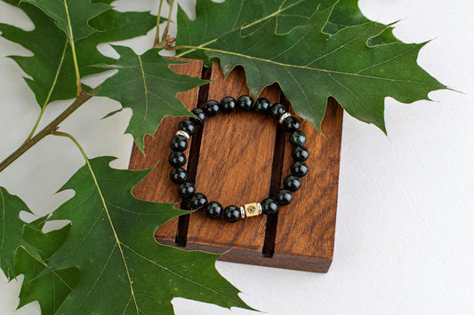 A bracelet with dark green beads and a gold-toned square charm rests on a wooden surface, surrounded by large green leaves.