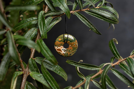A polished, donut-shaped pendant with green, orange and white patterns hangs from a black cord among dark green leaves.