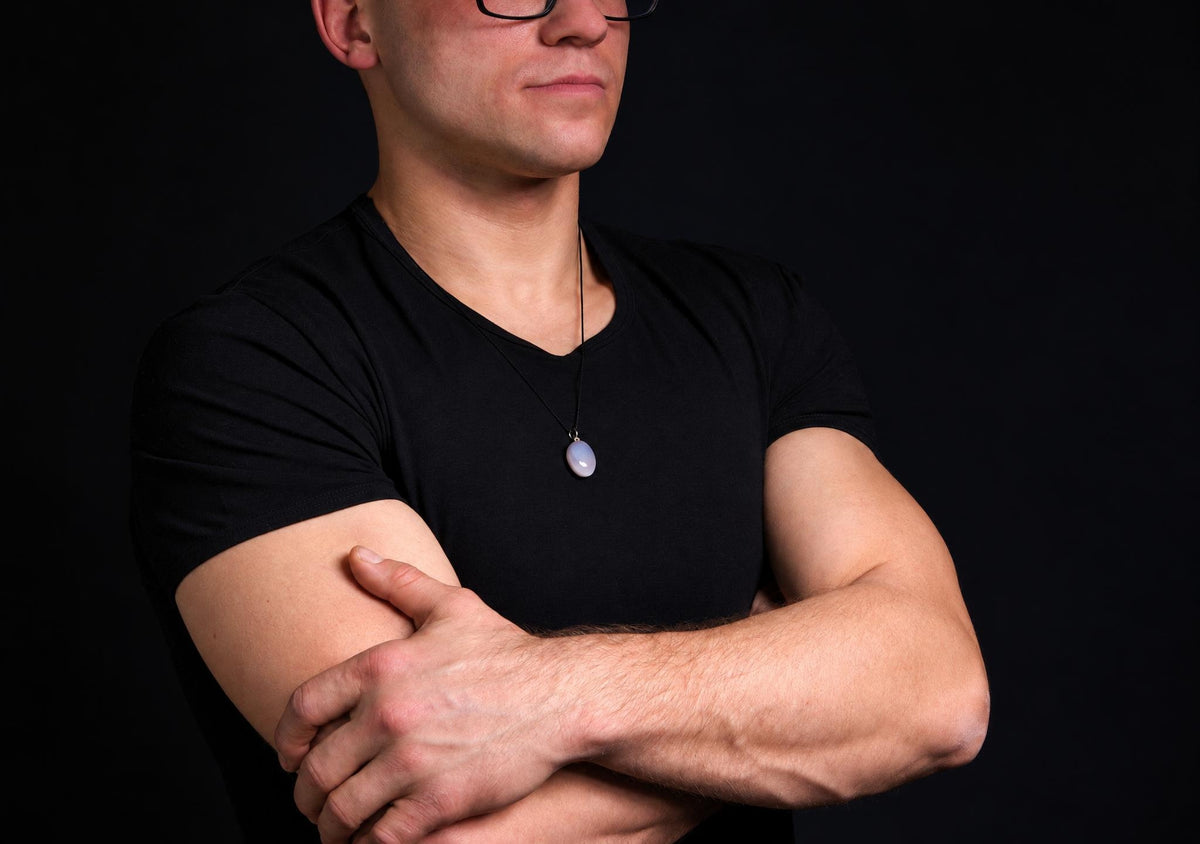 A man in a black t-shirt wears a pendant with an oval, pale grey stone on a dark cord against a dark background.