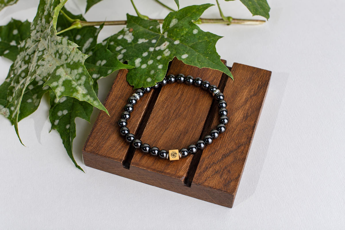 A bracelet with black spherical beads and a gold-coloured square charm rests on a dark wooden surface beside green leaves.
