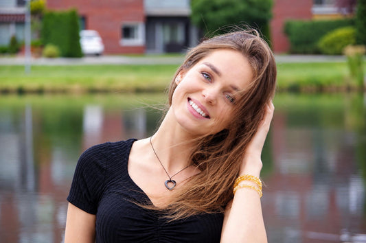 A woman wears a black top and a dark heart-shaped pendant necklace with yellow bracelets, smiling outdoors near water.