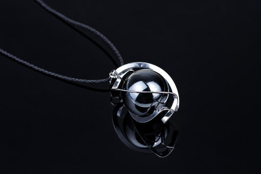 A silver-toned pendant with a dark, reflective sphere and curved metal frame hangs from a black cord against a dark surface.