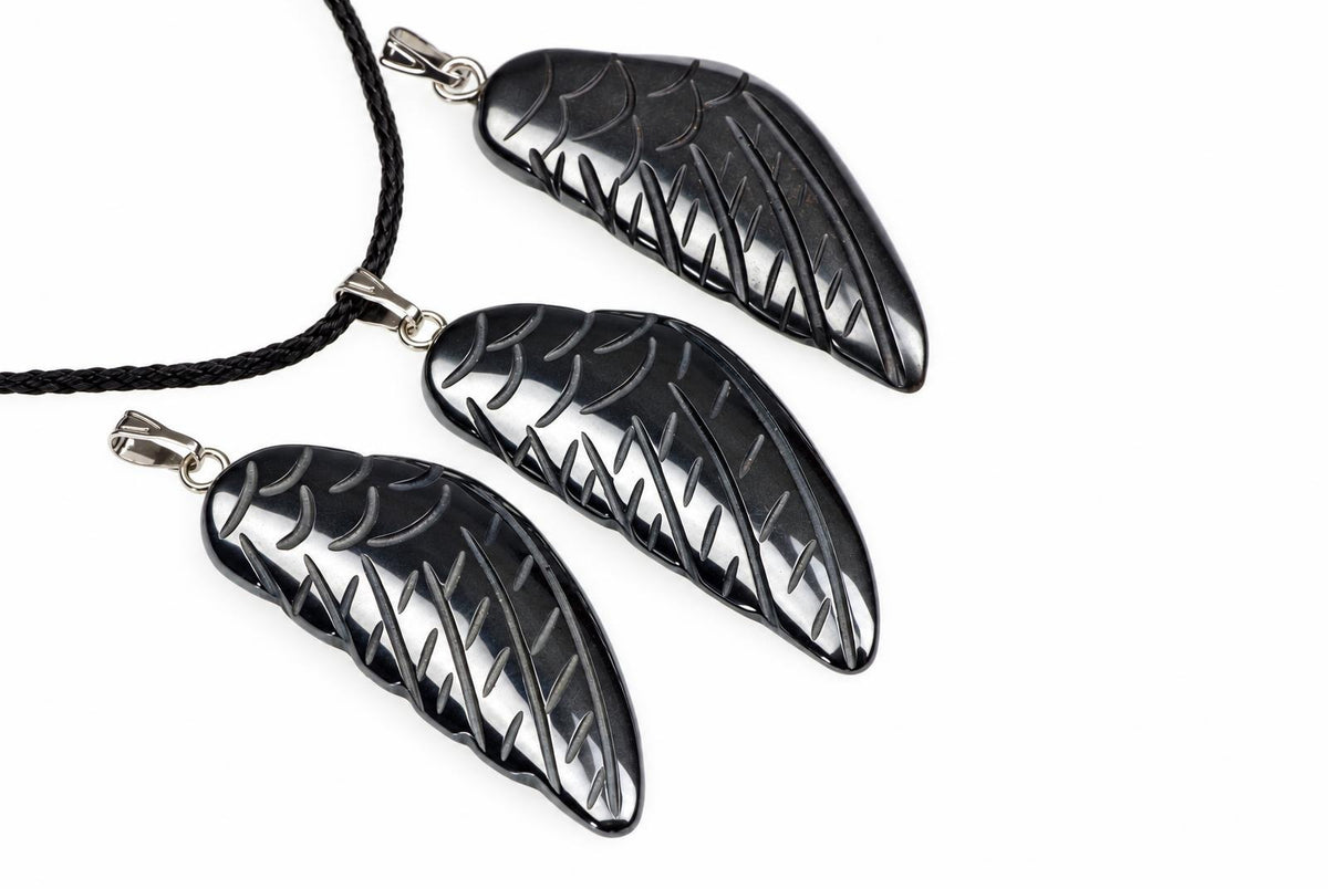 Dark polished wing-shaped hematite pendants with carved feather grooves and silver-tone loops are arranged with black cords against white.